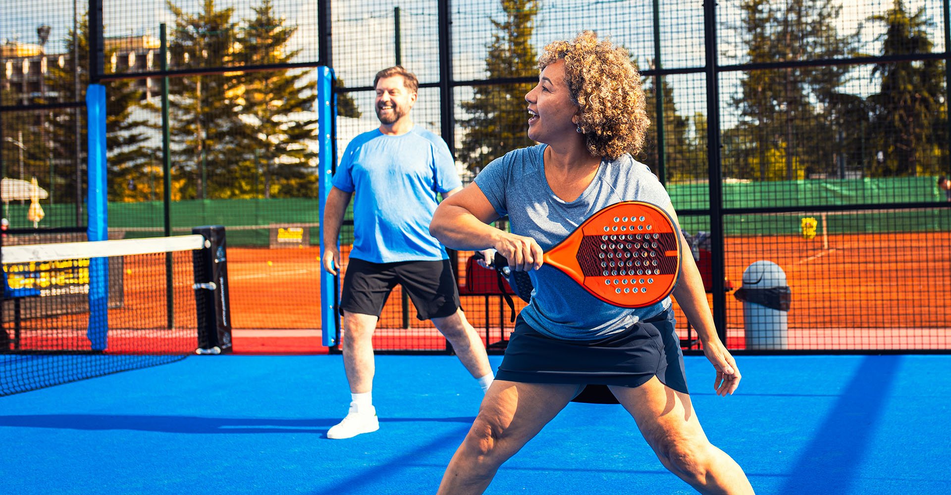 Mies ja nainen pelaavat padelia ulkokentällä. Heillä on yllään yhteensopivat asut, miehellä tummat shortsit ja vaaleansininen paita, naisella tumma lyhyt tennishame ja vaaleansininen paita. 