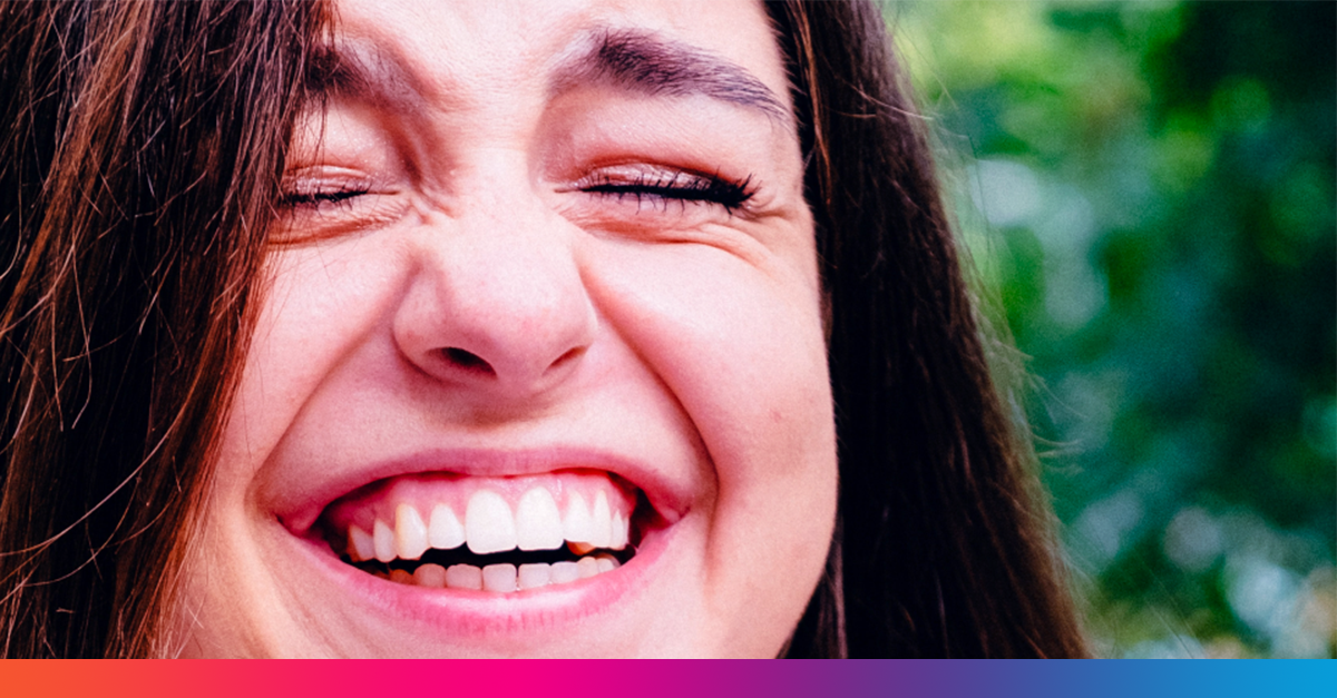 Nuori tummahiuksinen nainen nauraa, koska hänen valkoiset hampaansa ovat niin hyvin hoidetut ja olo sen mukainen.