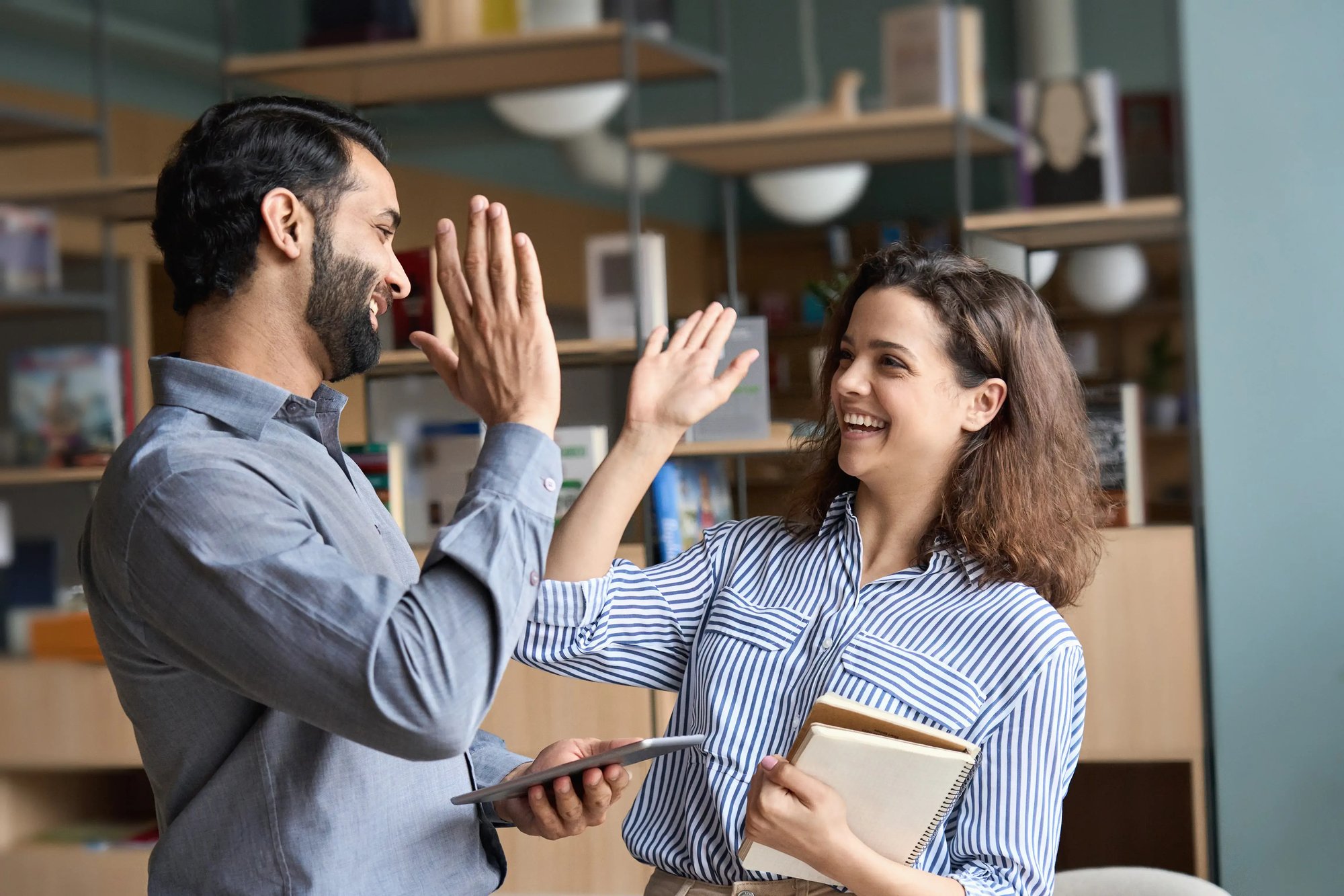 A man and a woman giving each other a high five indoors, with an office setting in the background. The individuals are happy and smiling at each other.
