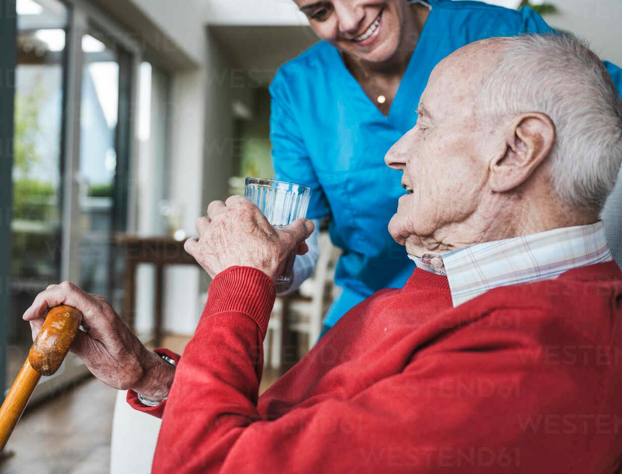 smiling-nurse-giving-water-to-senior-man-at-home-UUF29951