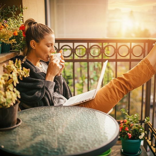 Nainen nauttimassa kahvia parvekkeella auringon paistaessa kannettava tietokone sylissä.
